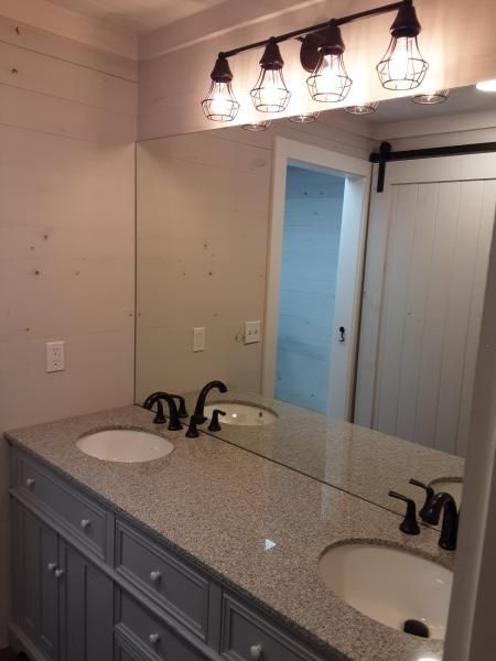 A bathroom vanity with grey cabinets, a speckled stone countertop, two sinks, bronze faucets, and a light fixture above.