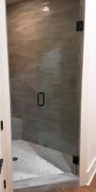 A glass shower door with a black handle and hinges, set against a tiled wall and a pebble-stone shower floor.