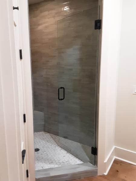 A walk-in shower with light-toned wood-look wall tiles, a white pebble floor, and a glass door with matte black hardware.