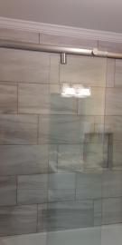 A sliding glass shower door with a stainless steel track mounted against a wall of light gray rectangular tiles.