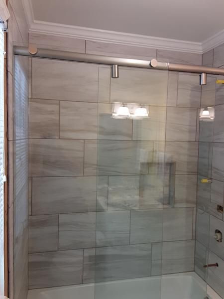 A glass sliding shower door with silver hardware installed over a bathtub against a grey tiled wall.