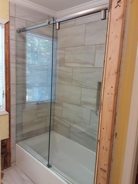 A frameless glass sliding shower door installed over a white bathtub in a tiled bathroom with unfinished wall framing.