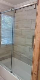 A frameless glass sliding shower door with brushed metal hardware installed over a bathtub with beige tiled walls.