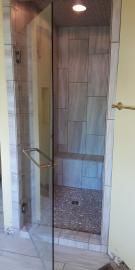 A glass shower door opens into a tiled walk-in shower with a pebble floor, bench, and recessed light.