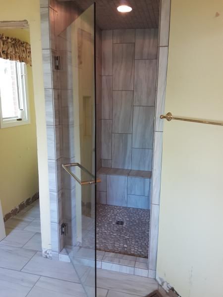 A walk-in shower with light gray tiled walls, a built-in bench, a pebble floor, and a glass door in a yellow bathroom.