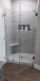 A walk-in shower with gray tiled walls, a corner seat, a recessed shelf, and a glass door in a bathroom with wood floors.