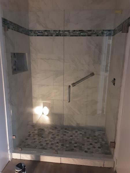A walk-in shower with light marble-style tiles, a pebble-stone floor, a glass door, and a horizontal grab bar inside.