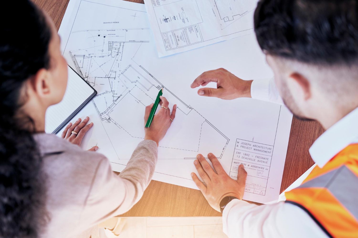 Two people examine and point to architectural blueprints on a table, one wearing a high-visibility safety vest.