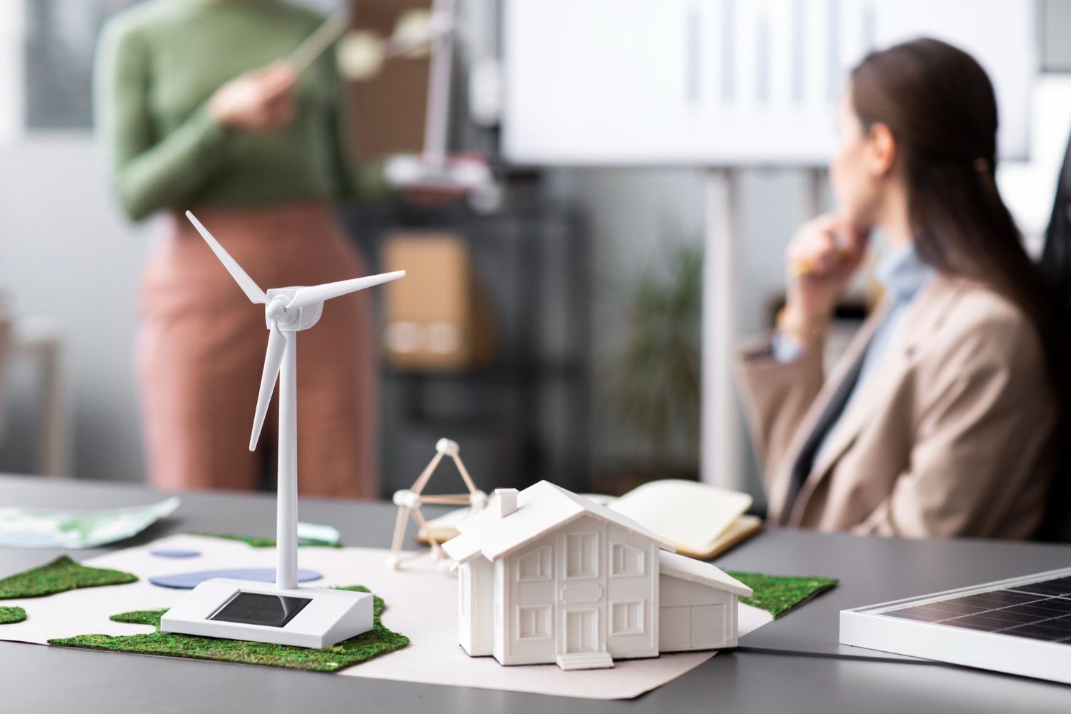 A miniature wind turbine and house model sit on a desk in front of two people in a business meeting.