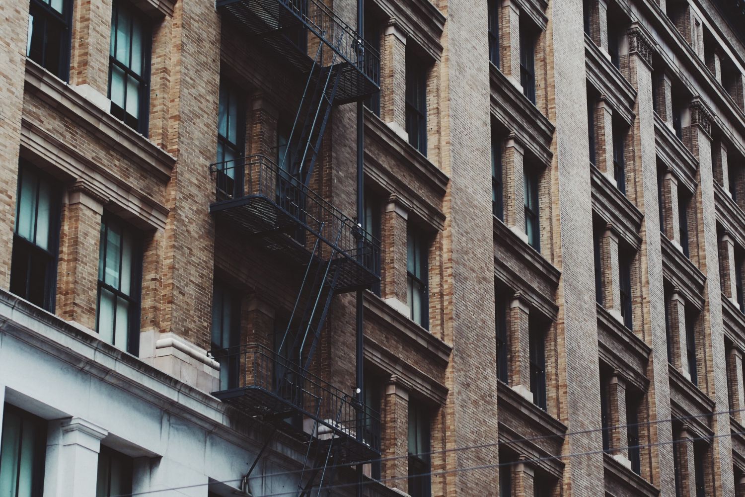 A low-angle view of a brick apartment building in a city, featuring classic black metal fire escapes and grid windows.
