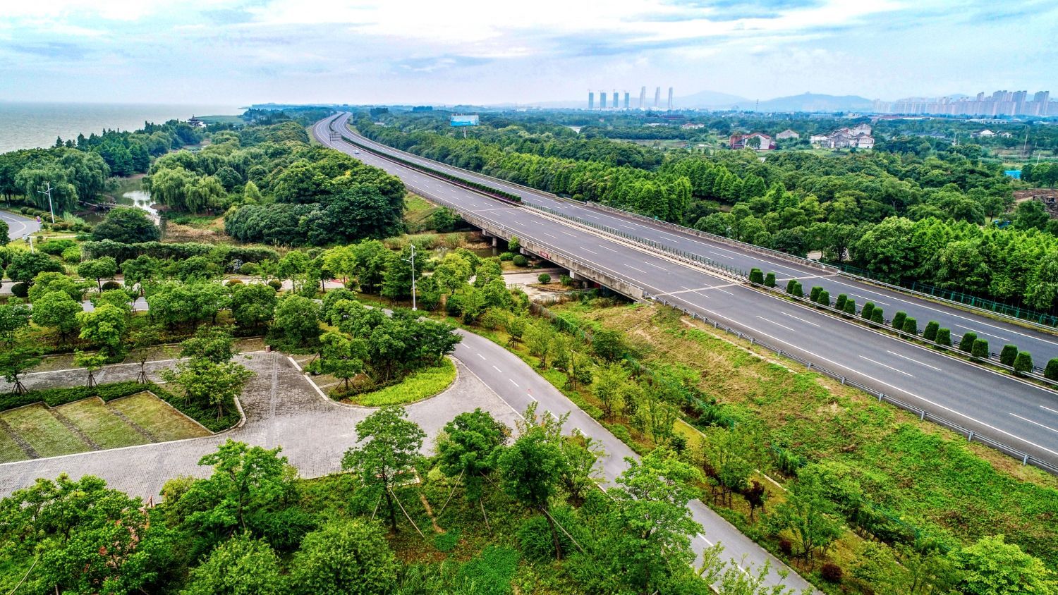 An elevated multi-lane highway curves through a lush, green landscape under a cloudy sky, near a body of water.