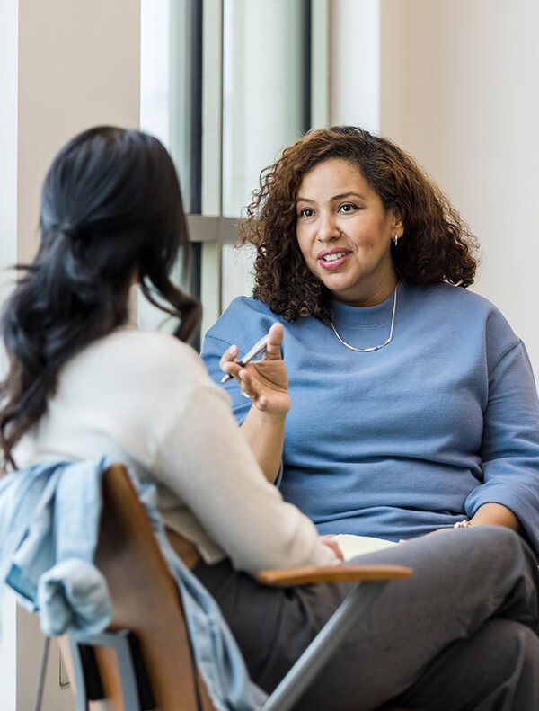 Two people in conversation, one holding pen and talking, in a brightly lit setting.