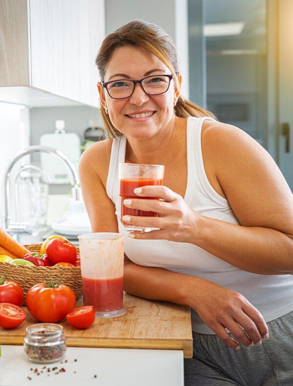 Woman in glasses holding juice in kitchen, smiling, vegetables nearby.