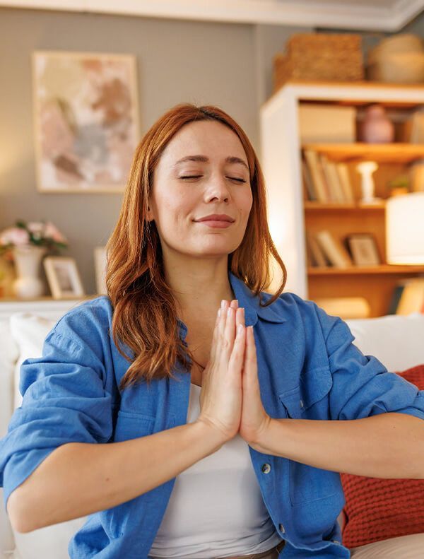 Woman with red hair, eyes closed, hands in prayer pose, smiling, in a living room.