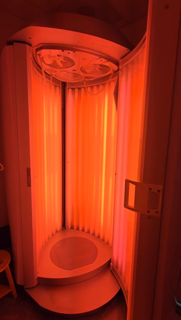 Woman wearing eye protection lies under red light in a spa. She is wrapped in a white robe, relaxing with eyes closed.
