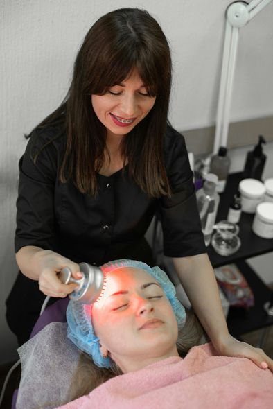 A woman administers red light therapy to another woman's face in a salon setting. The recipient wears a hair net and appears relaxed.