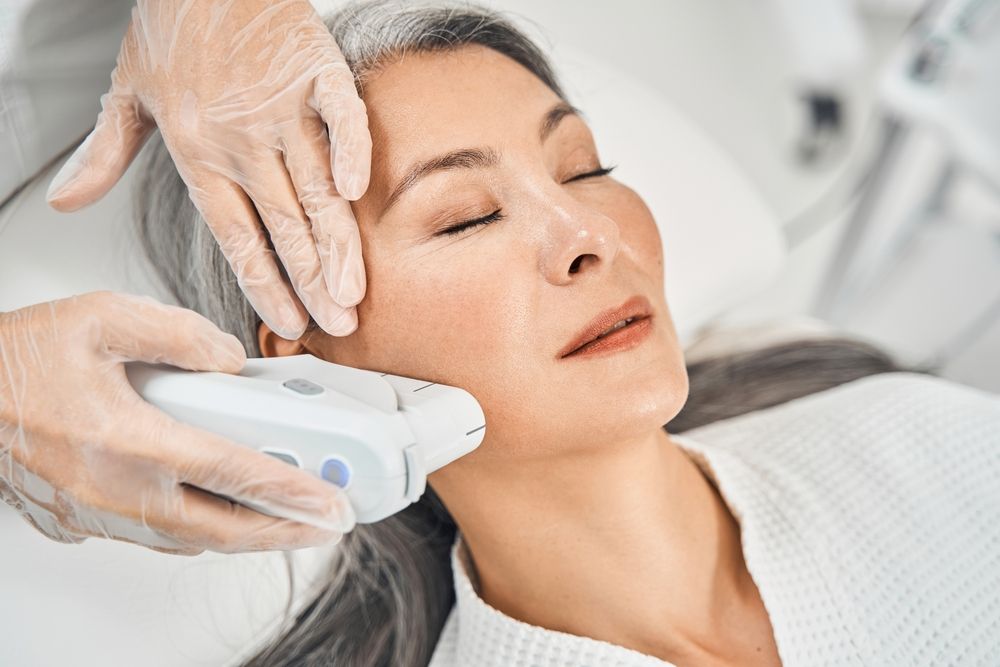 Aesthetician performing a facial treatment on a woman. The woman has her eyes closed and is wearing a white robe.