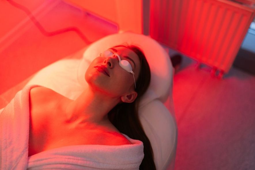 Woman wearing eye protection lies under red light in a spa. She is wrapped in a white robe, relaxing with eyes closed.