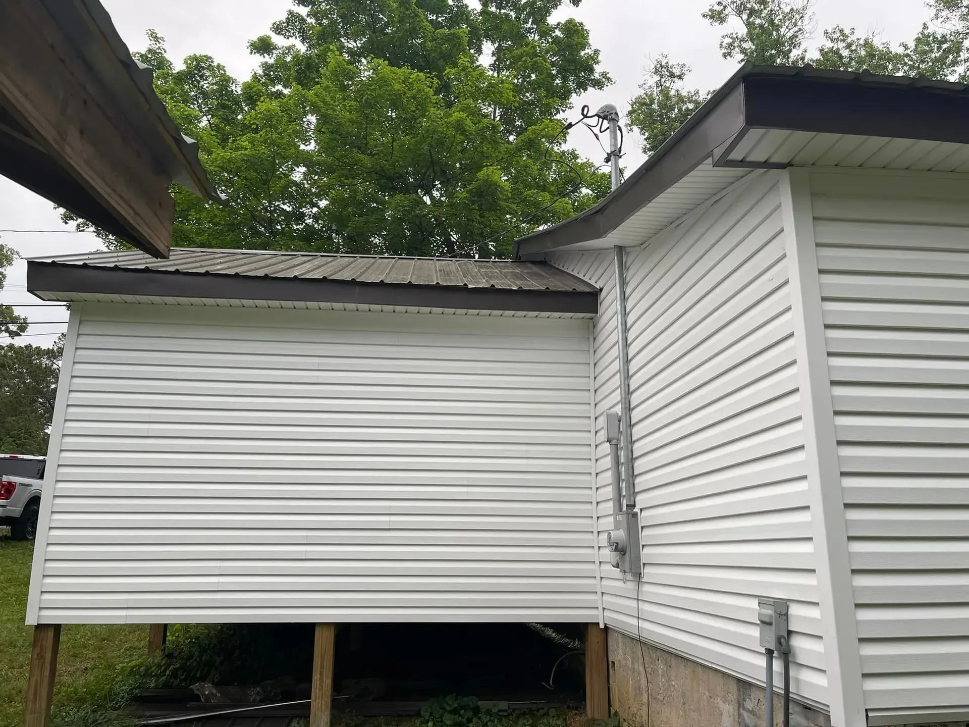 White vinyl-sided building with a dark brown roof, surrounded by trees and an exterior power panel.