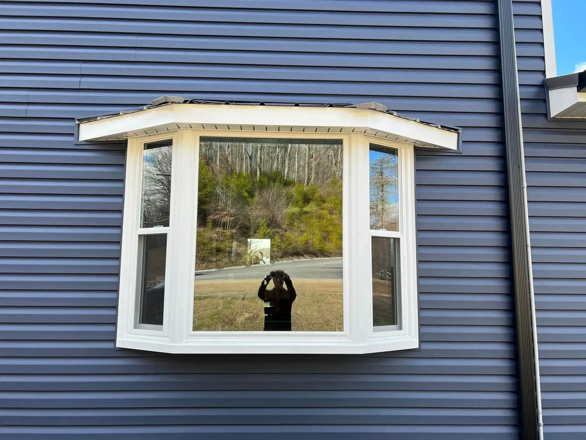 Bay window on a gray-sided house, reflecting a person taking a photo and a road with trees.