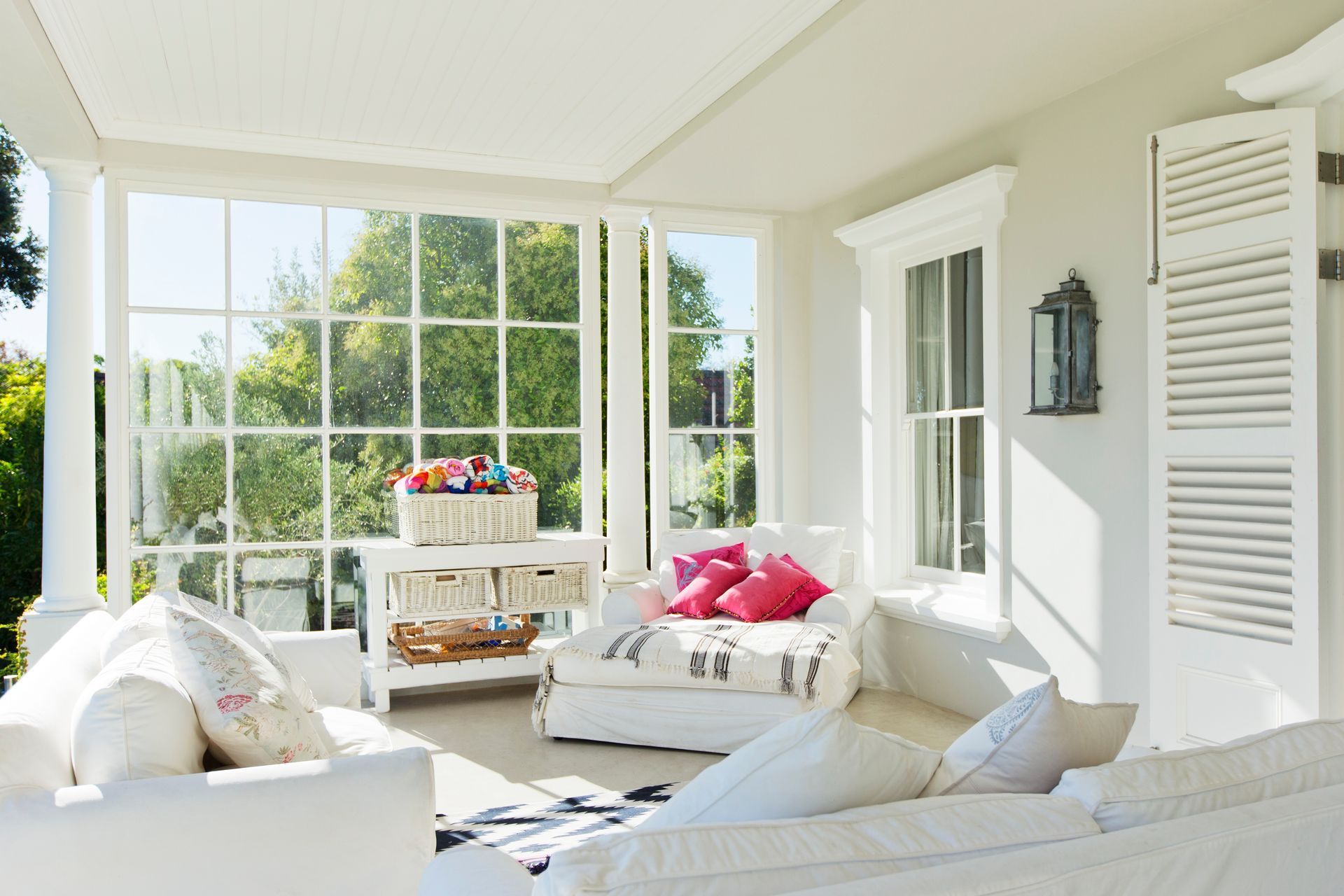 Sunlit white porch with large windows, white furniture, and colorful pillows.