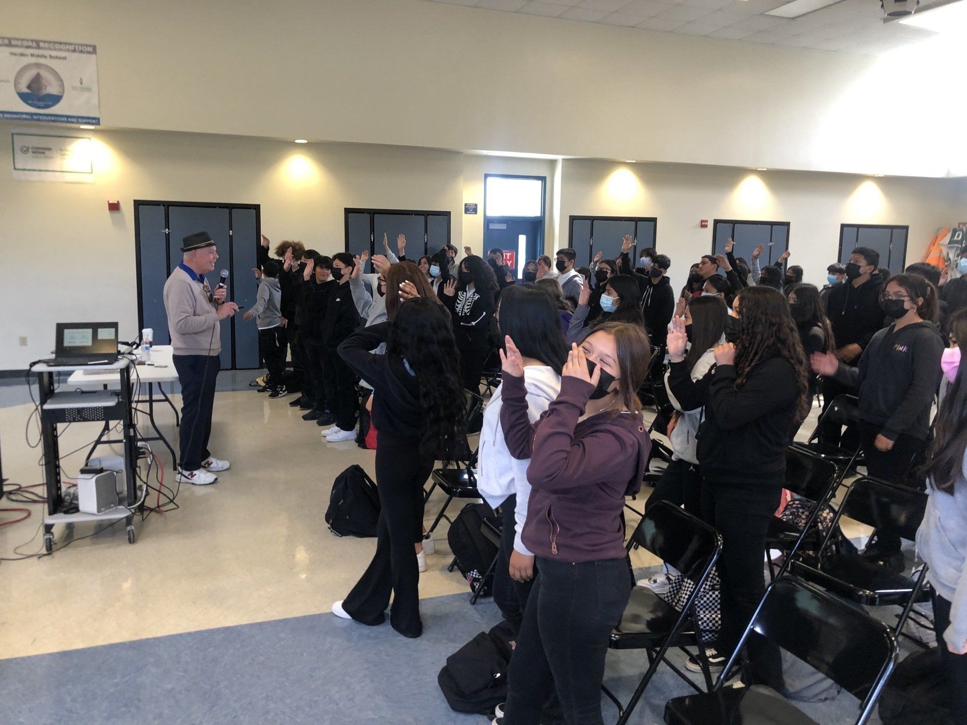 A man speaks to a group of students in a room. Students raise their hands.
