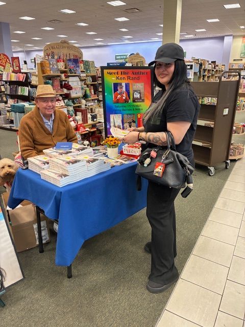 Man signing books at a table in a bookstore; woman with purse looks on. A dog is visible.