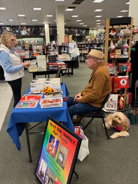Author at table in bookstore, speaking to a woman. A dog sits nearby, watching.