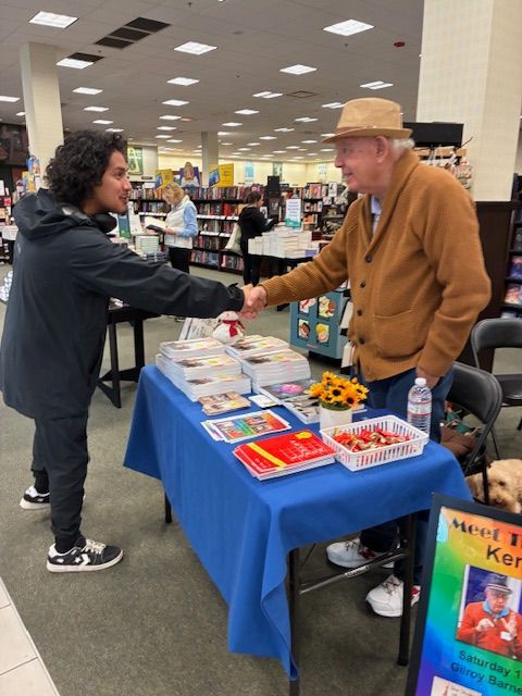 Man shaking hands with another man at a bookstore table, books, and colorful sign.