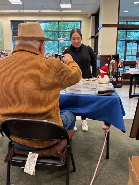 Person in brown hat and sweater at table with books; woman behind table smiling. Bookstore setting.