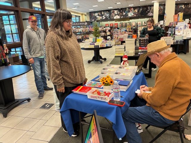 A person signing books at a table in a bookstore. Another person watches nearby.