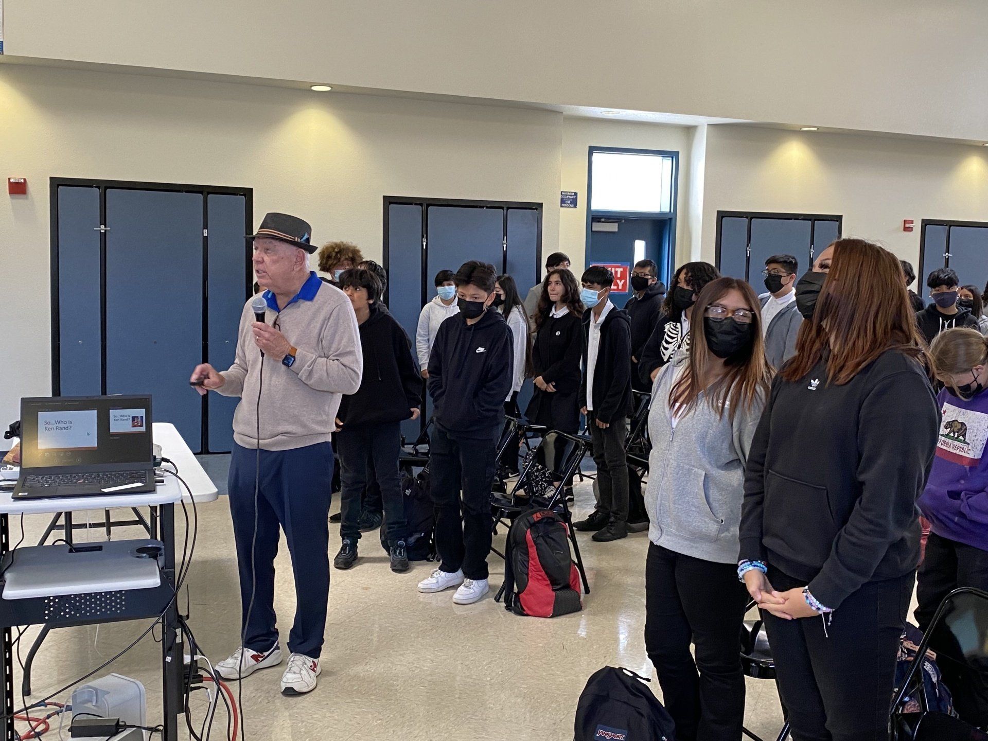 A man in a hat presents to a group of students in a large room.