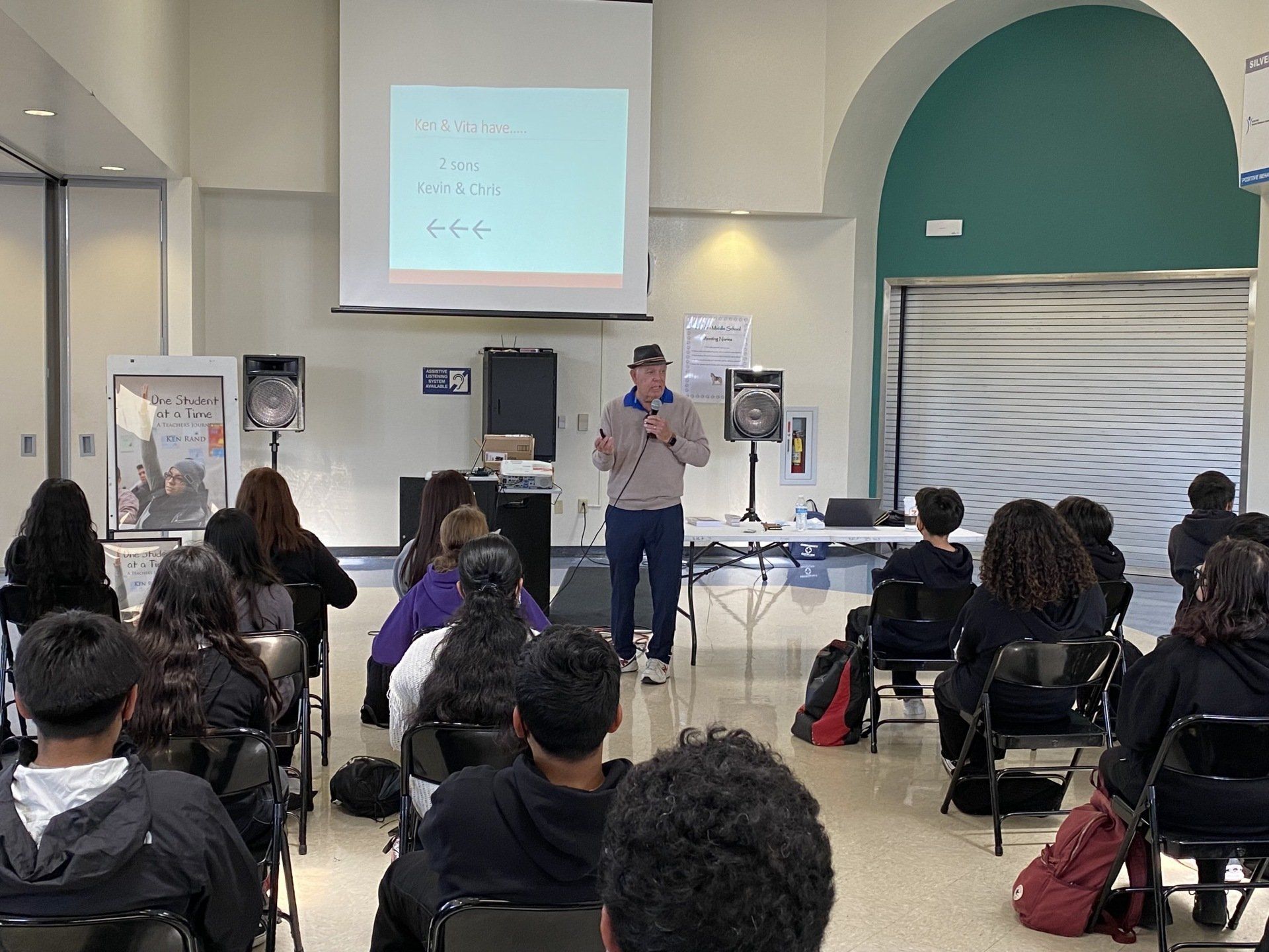 Man presenting to a group of students. A projector displays equations; students seated in chairs, a room with an arch.