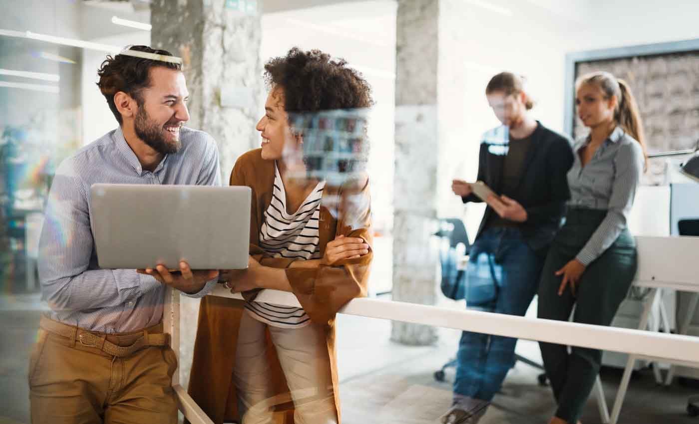 A man is holding a laptop and talking to a woman in an office.