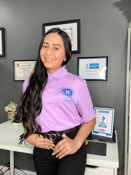 A person with long dark hair smiles in a light purple polo shirt and dark pants, standing in an office with framed awards.