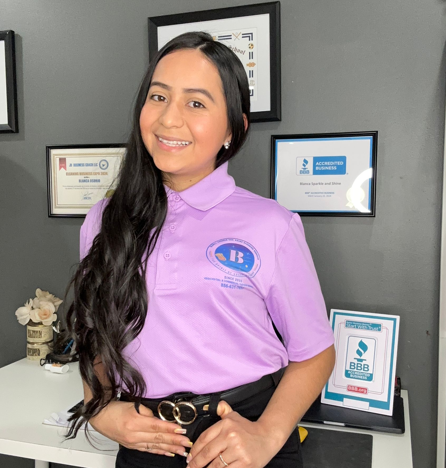A person with long dark hair smiles in a light purple polo shirt and dark pants, standing in an office with framed awards.