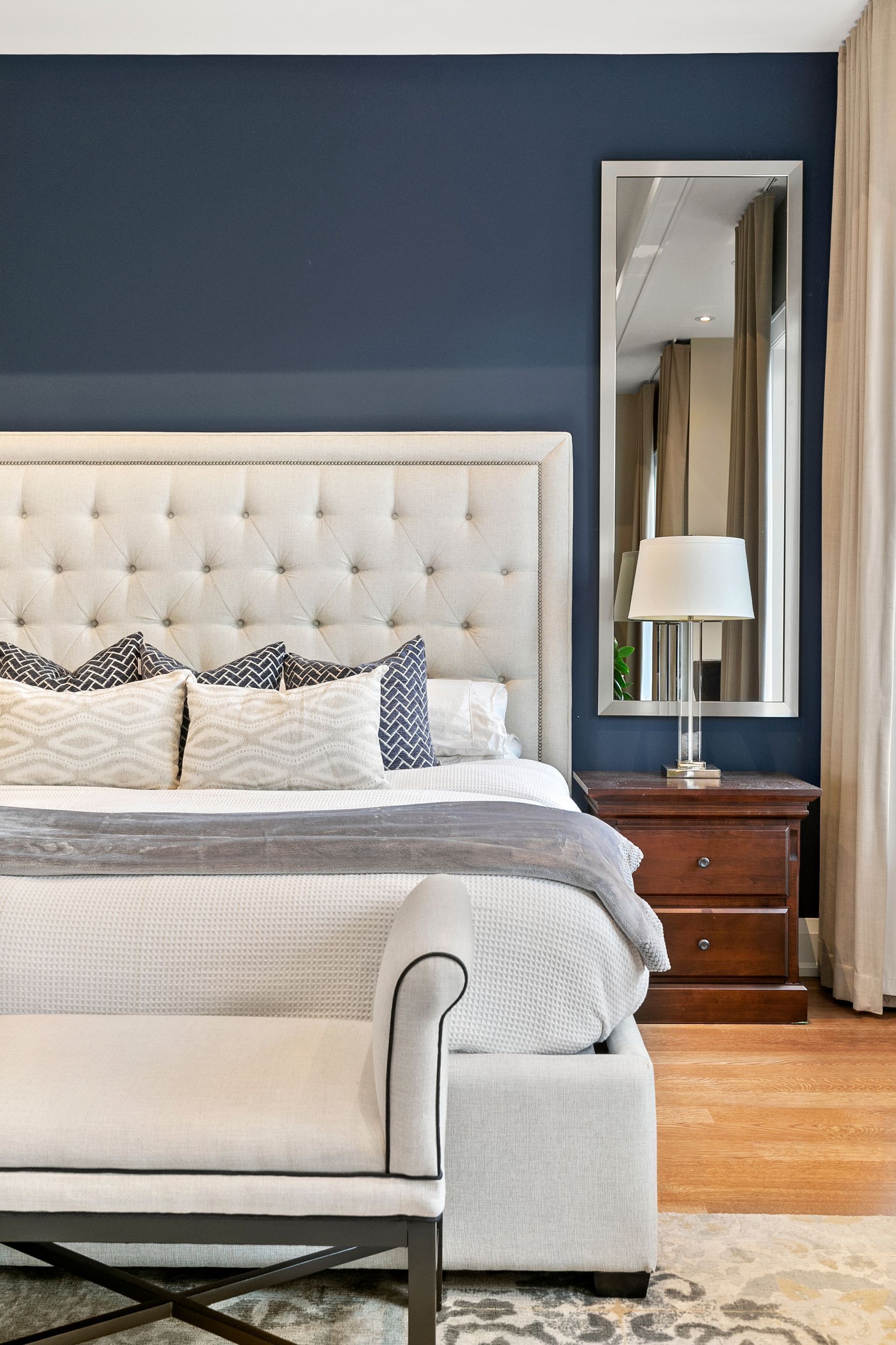 Bedroom with a tufted headboard, dark blue wall, and tall mirror. Includes a nightstand, bed bench, and cream-colored curtains.