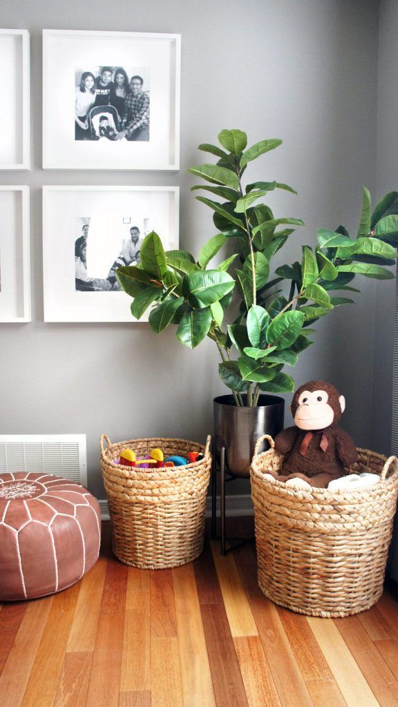 A brown leather ottoman, wicker baskets, and a leafy plant beside a gray wall with family photos.