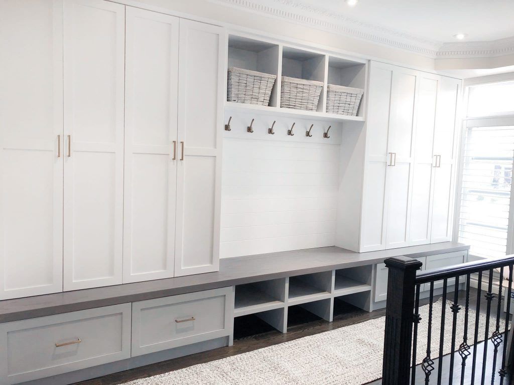 White built-in storage with upper cabinets, baskets, hooks, bench, and lower drawers. Gray accent colors.