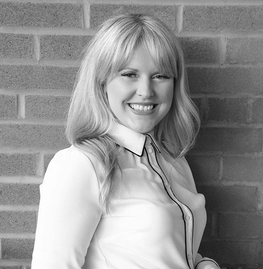 Blonde woman smiles at the camera, leaning against a brick wall; wearing a white shirt.