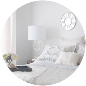 Bedroom with white bedding, lamp, tray with flowers, and round wall mirror.