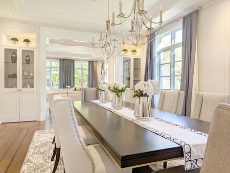 Elegant dining room with long dark table, white chairs, chandelier, and large windows with gray curtains.
