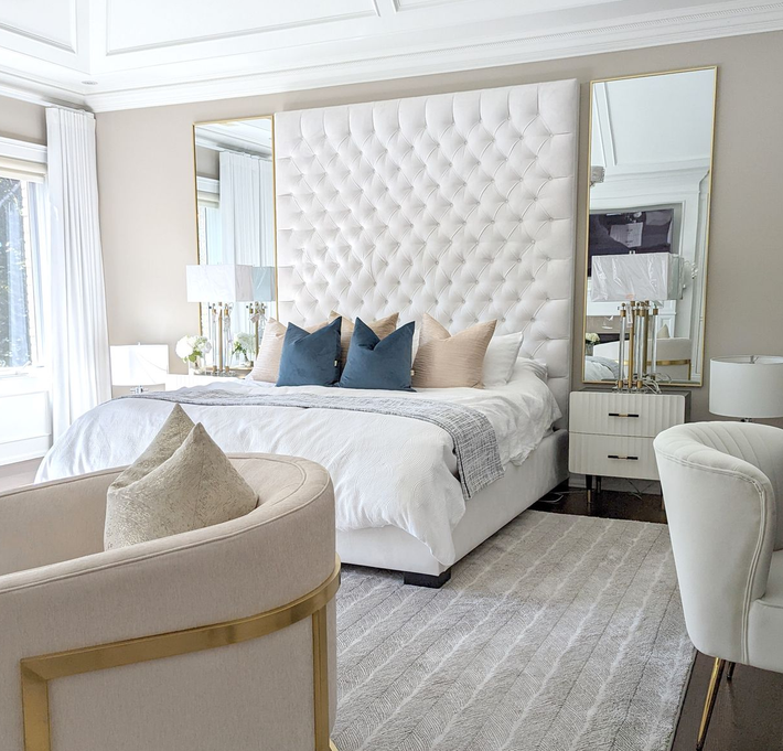 Luxurious bedroom with a tufted white headboard, mirrors, and a neutral color scheme.
