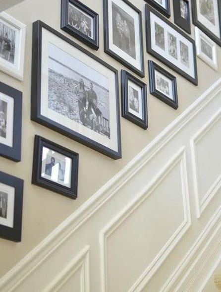 Black and white framed photos arranged on a beige wall above white paneling along a staircase.