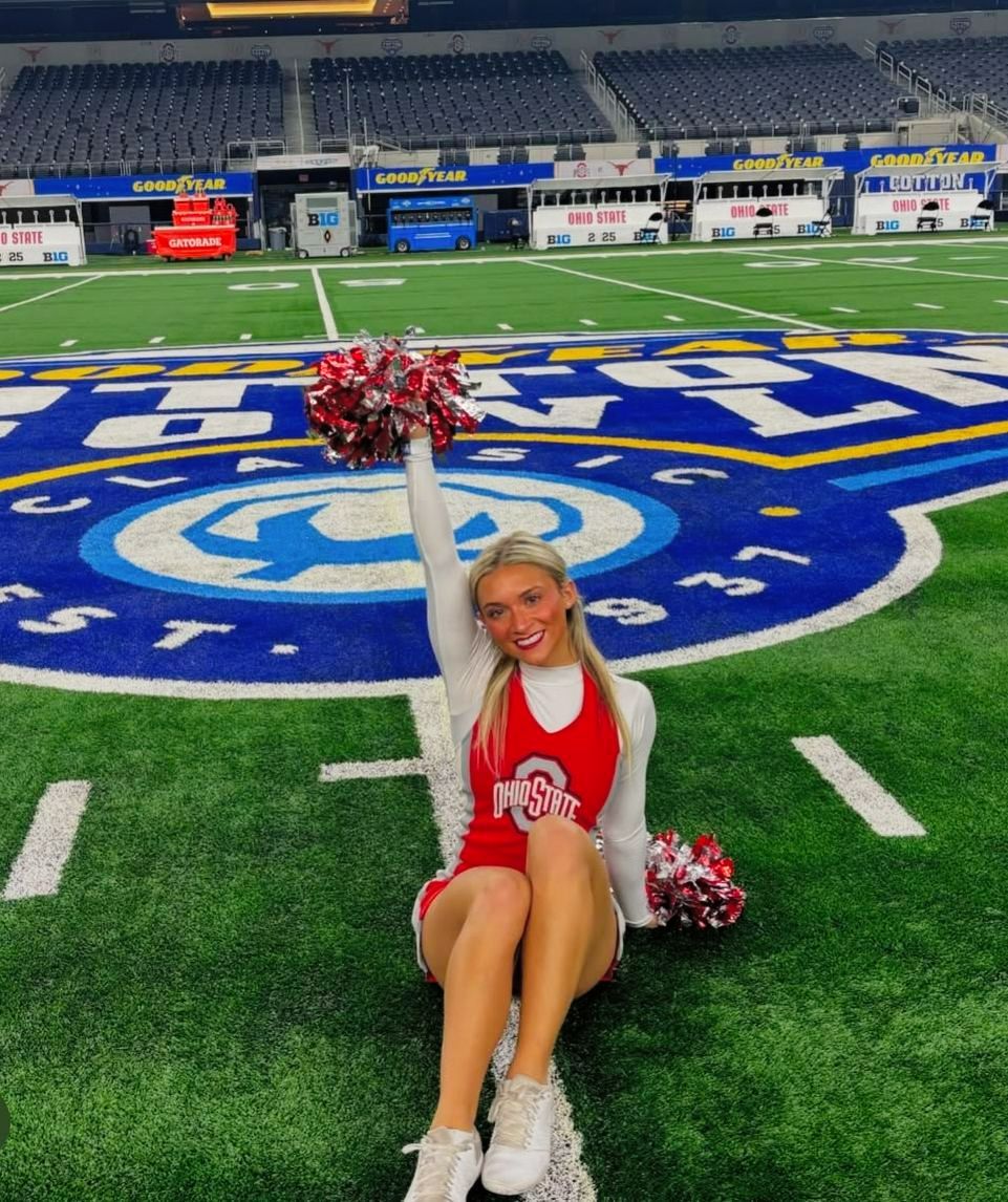 A cheerleader is sitting on a football field holding pom poms.