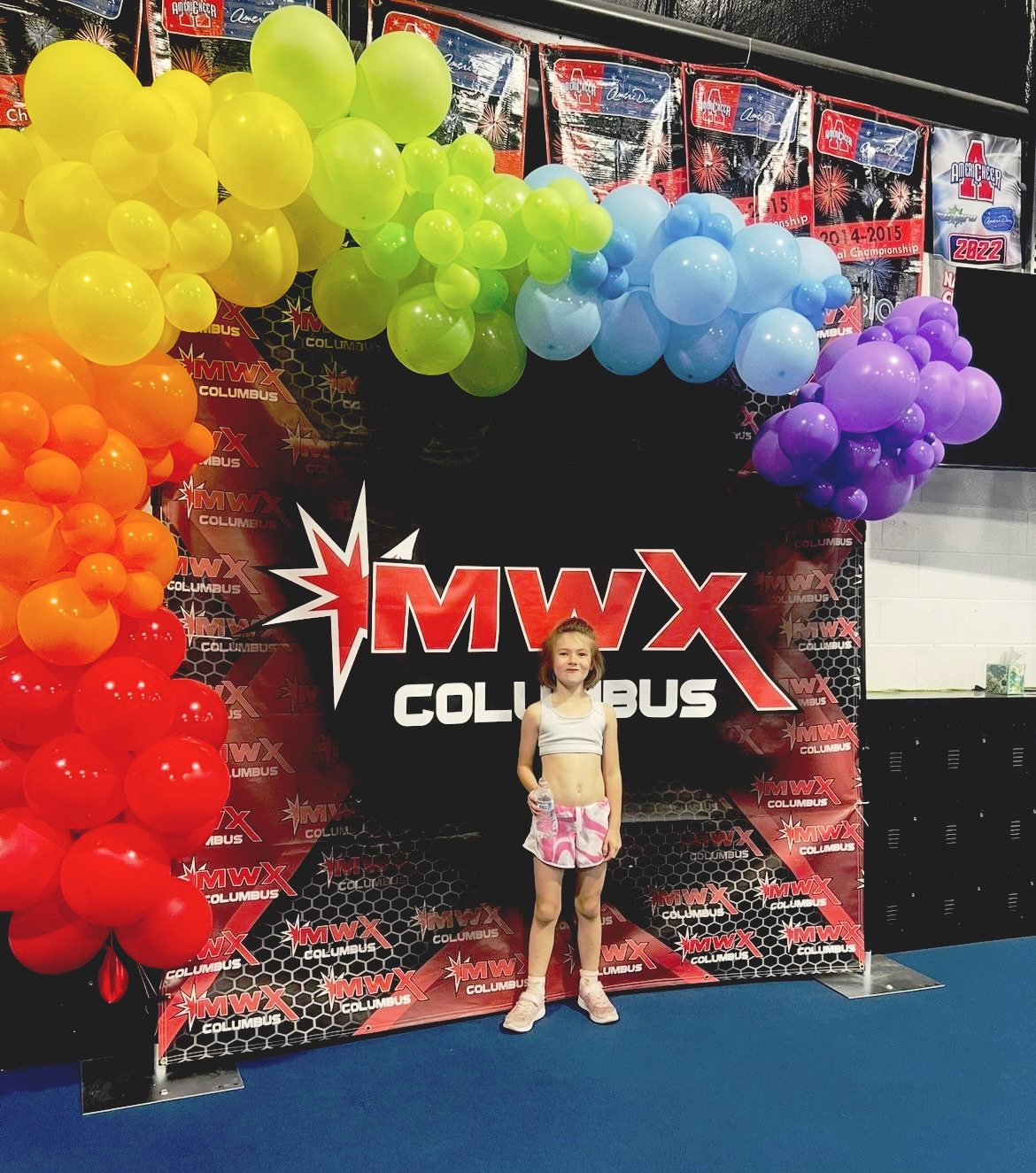 Girl in cheerleading outfit poses under rainbow balloon arch at MWX Columbus event.