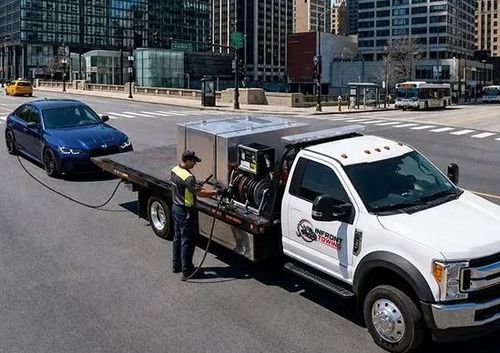 Tow truck towing a blue car on a city street, with a worker standing beside it.