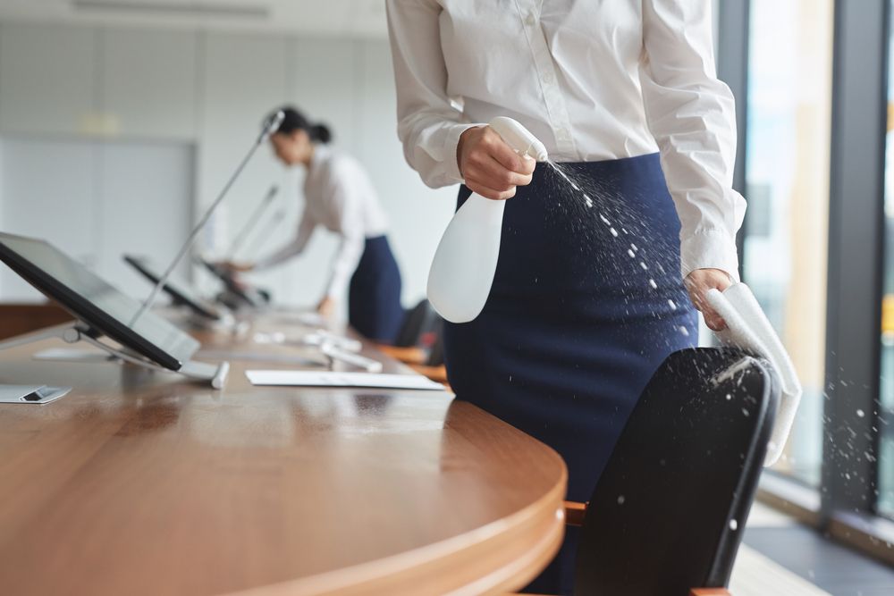 Woman spraying disinfectant on chair in a modern office, another cleaning in the background.