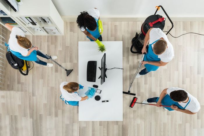 Cleaners in blue aprons cleaning an office, using vacuums, spray bottles, and cloths.