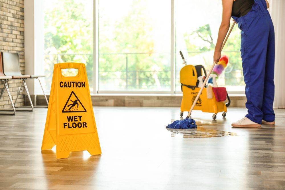 Yellow wet floor sign near a person mopping a light wood floor, in a bright room.
